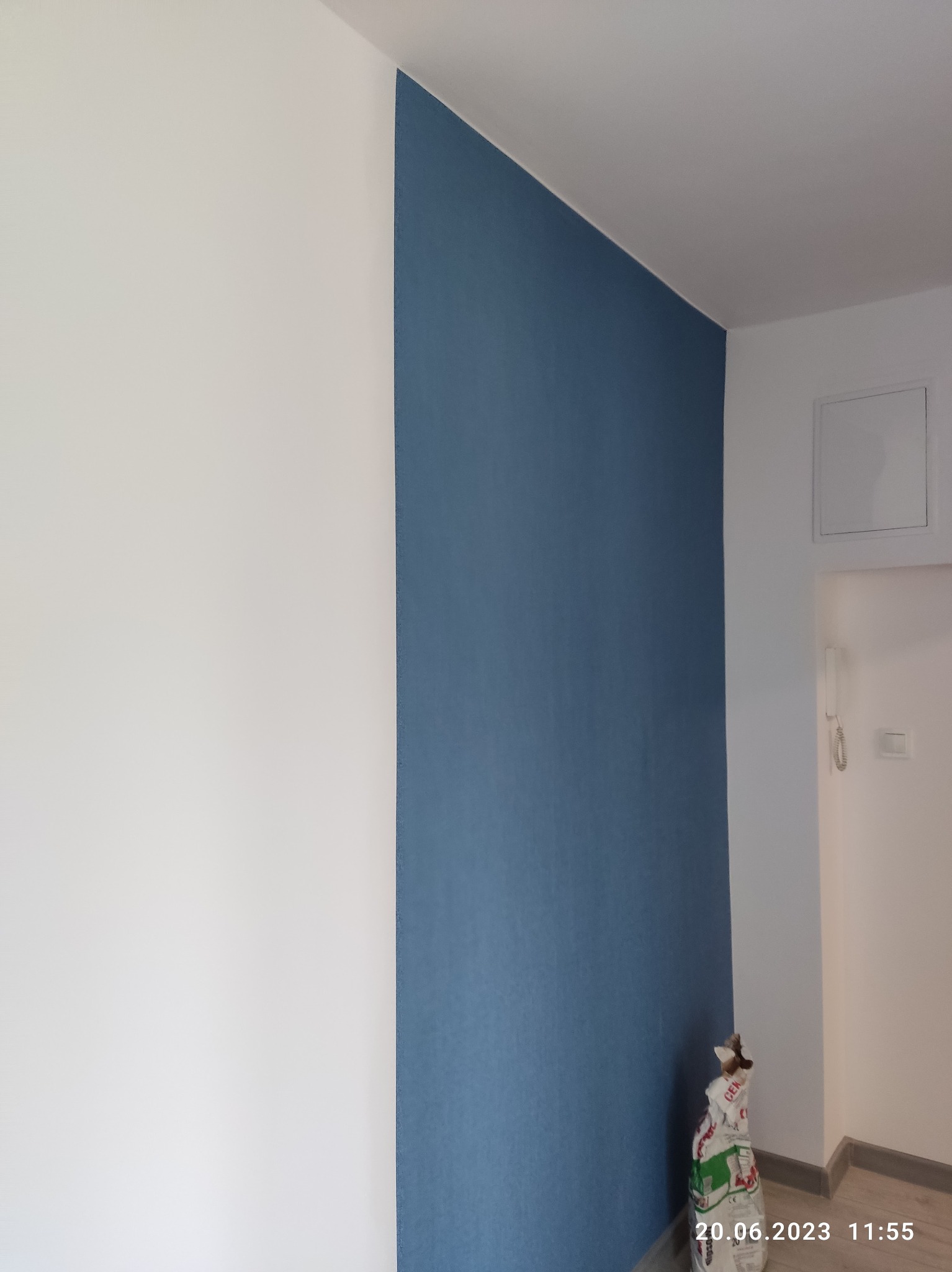 Pomieszczenie po remoncie: biała ściana sąsiaduje z fragmentem ściany pomalowanej na niebiesko, w rogu stoi worek po materiałach budowlanych.