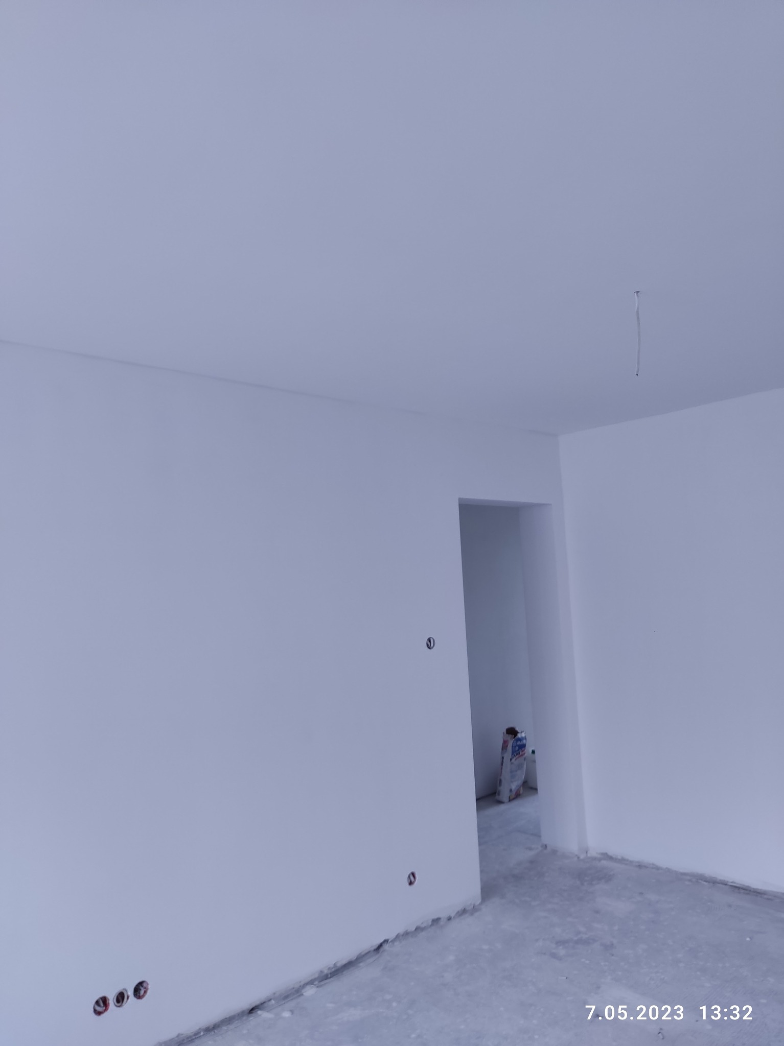 Pomalowane na biało wnętrze pokoju z widocznymi gniazdkami elektrycznymi, surową podłogą i workiem z materiałem budowlanym w przejściu do kolejnego pomieszczenia.