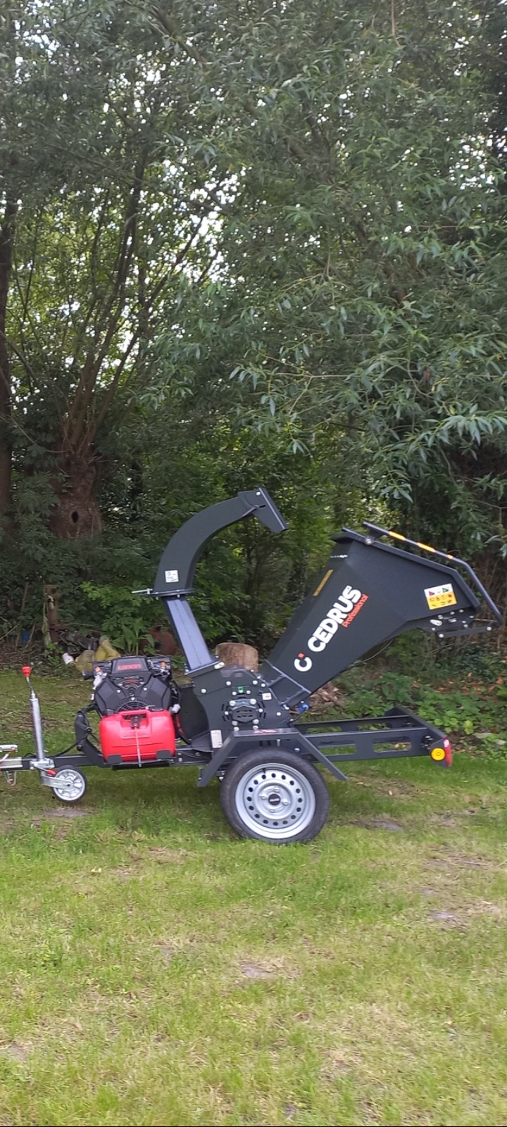 Rębak do gałęzi Cedrus Professional na trawie, gotowy do pracy. Maszyna z silnikiem spalinowym, z dyszą wylotową i kołami, tło stanowią drzewa.