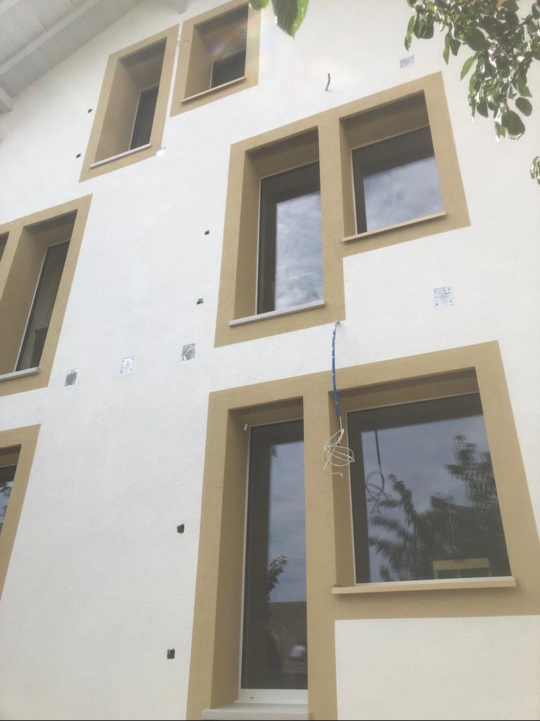 Elewacja budynku w trakcie prac: widoczne otwory po kołkach, fragmenty siatki z klejem, okna z brązowymi obramowaniami i tynk w kolorze kremowym.