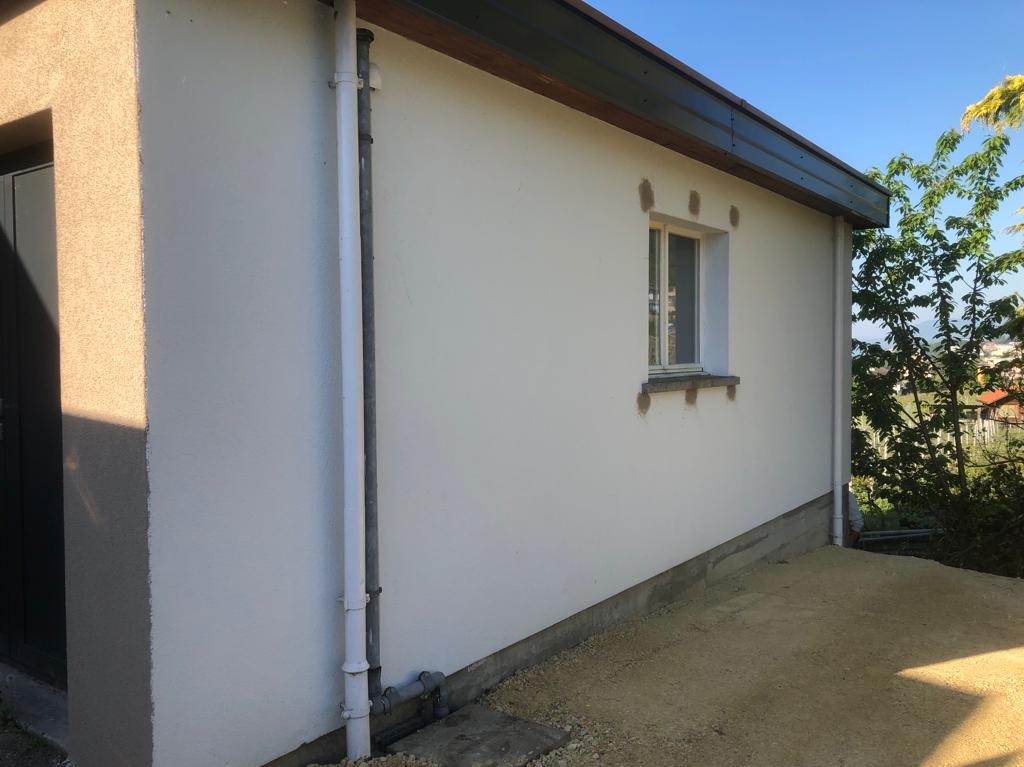 Ściana budynku po tynkowaniu z widocznymi łatami wokół okna, rura spustowa i fragment zieleni w tle, ukazująca etap prac elewacyjnych.