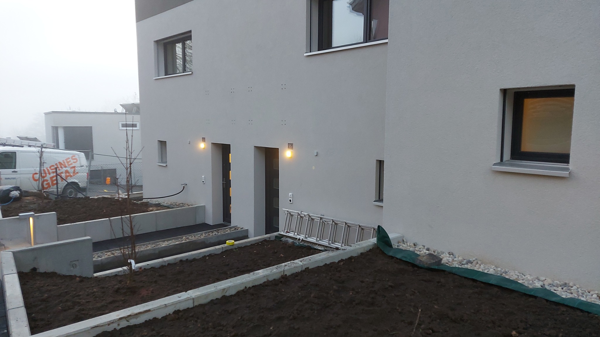 Nowoczesny budynek z szarą elewacją w trakcie prac wykończeniowych ogrodu, drabina oparta o ścianę. W tle widoczny samochód dostawczy.