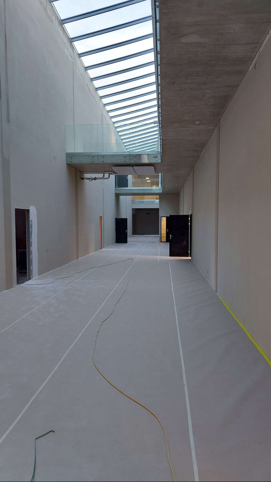 Wnętrze w trakcie wykończenia: szpachlowane ściany, surowy beton, przeszklony sufit i widoczne kable na podłodze. Otwory drzwiowe bez ościeżnic czekają na montaż.