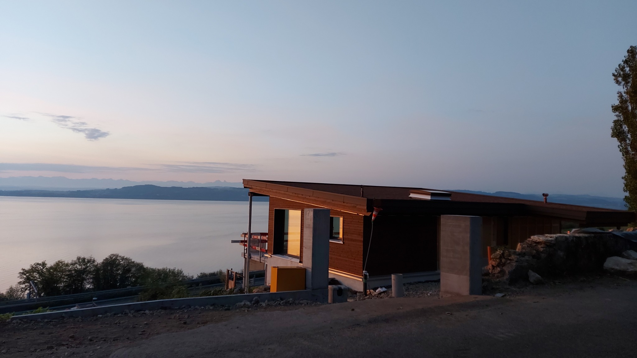 Nowoczesny dom z drewnianą elewacją nad jeziorem, w trakcie budowy. Widoczne rusztowania i elementy konstrukcyjne. Zachód słońca odbija się w oknach.