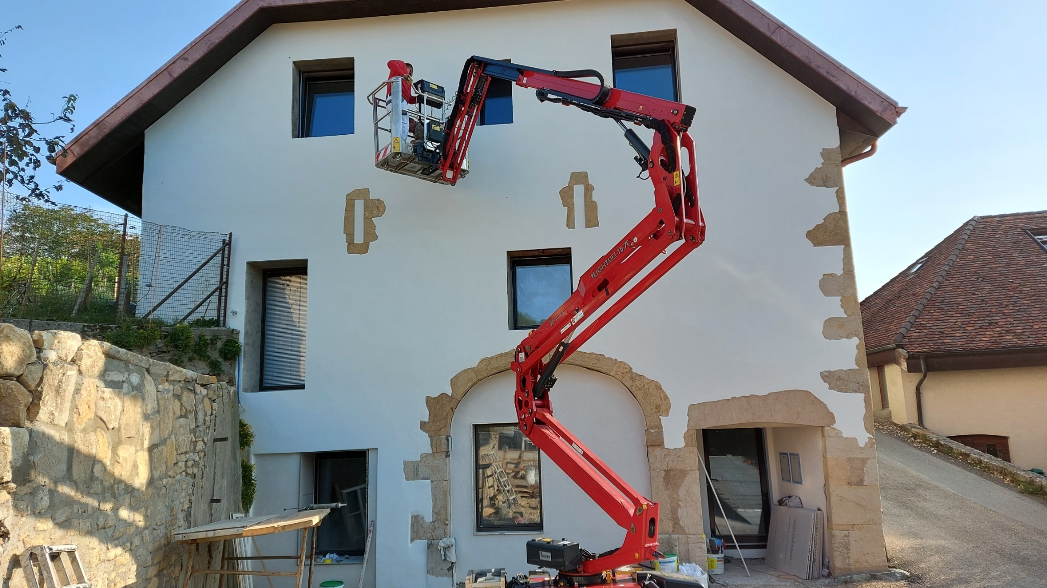 Prace na elewacji budynku z wykorzystaniem podnośnika koszowego. Częściowo usunięty stary tynk, przygotowanie do nałożenia nowej warstwy. Widoczne narzędzia pracy.