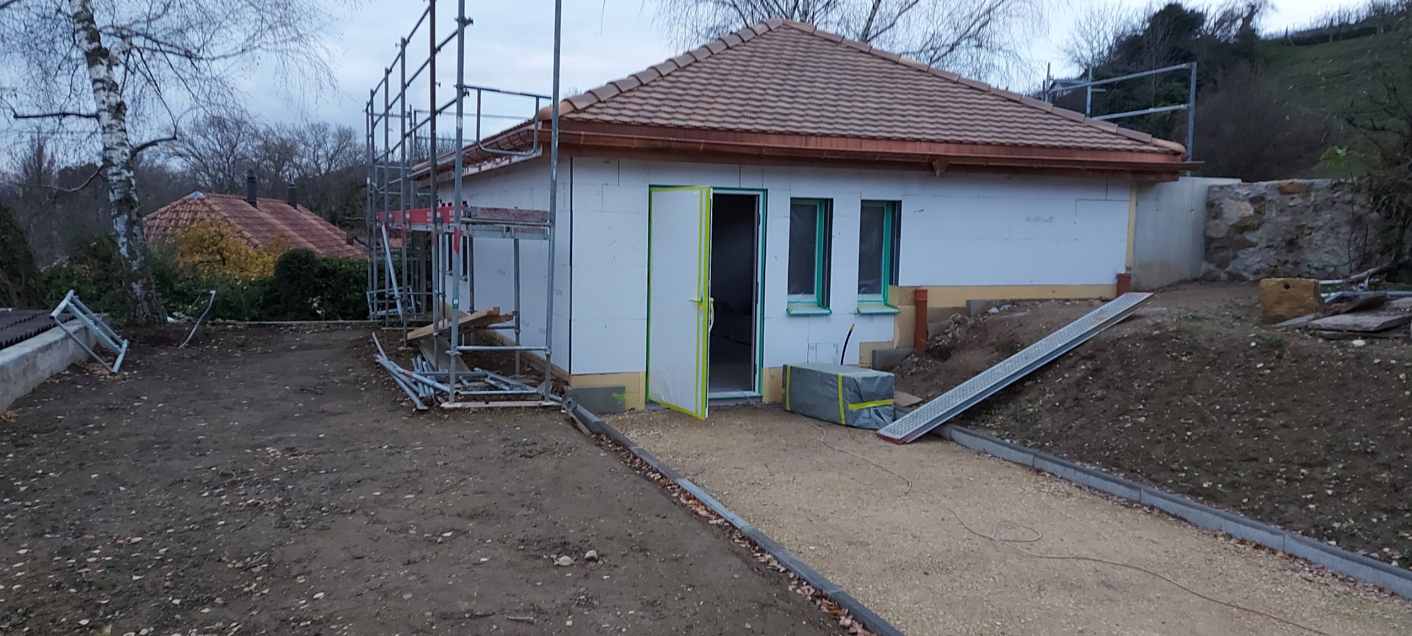 Budynek w trakcie ocieplania elewacji, widoczne rusztowania i materiały izolacyjne. Otwarta brama wejściowa, teren wokół w trakcie prac wykończeniowych.