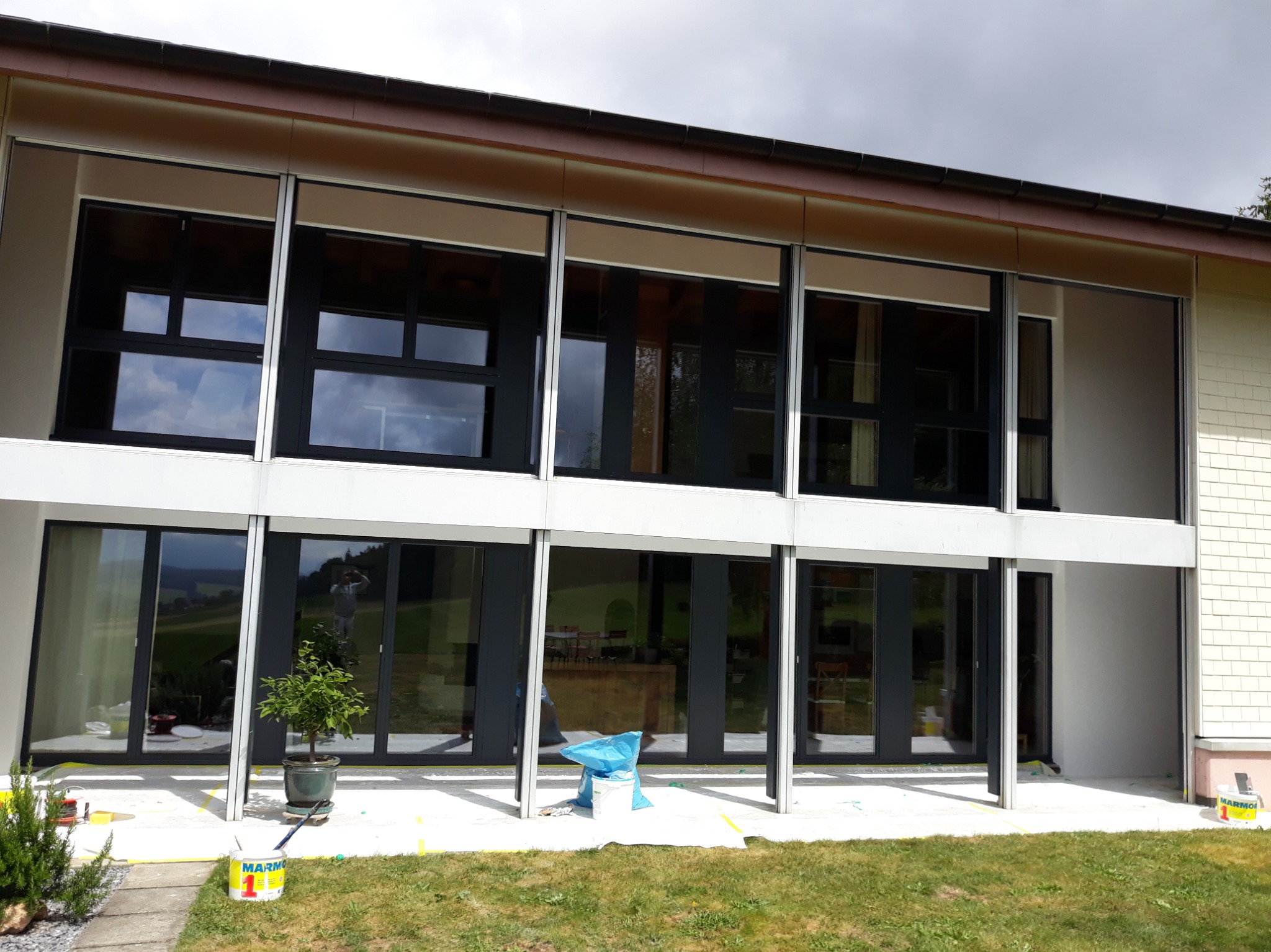 Elewacja domu z dużymi oknami w trakcie malowania. Widoczne farby, zabezpieczenia. Częściowo pomalowana ściana, na trawniku wiadro z farbą.