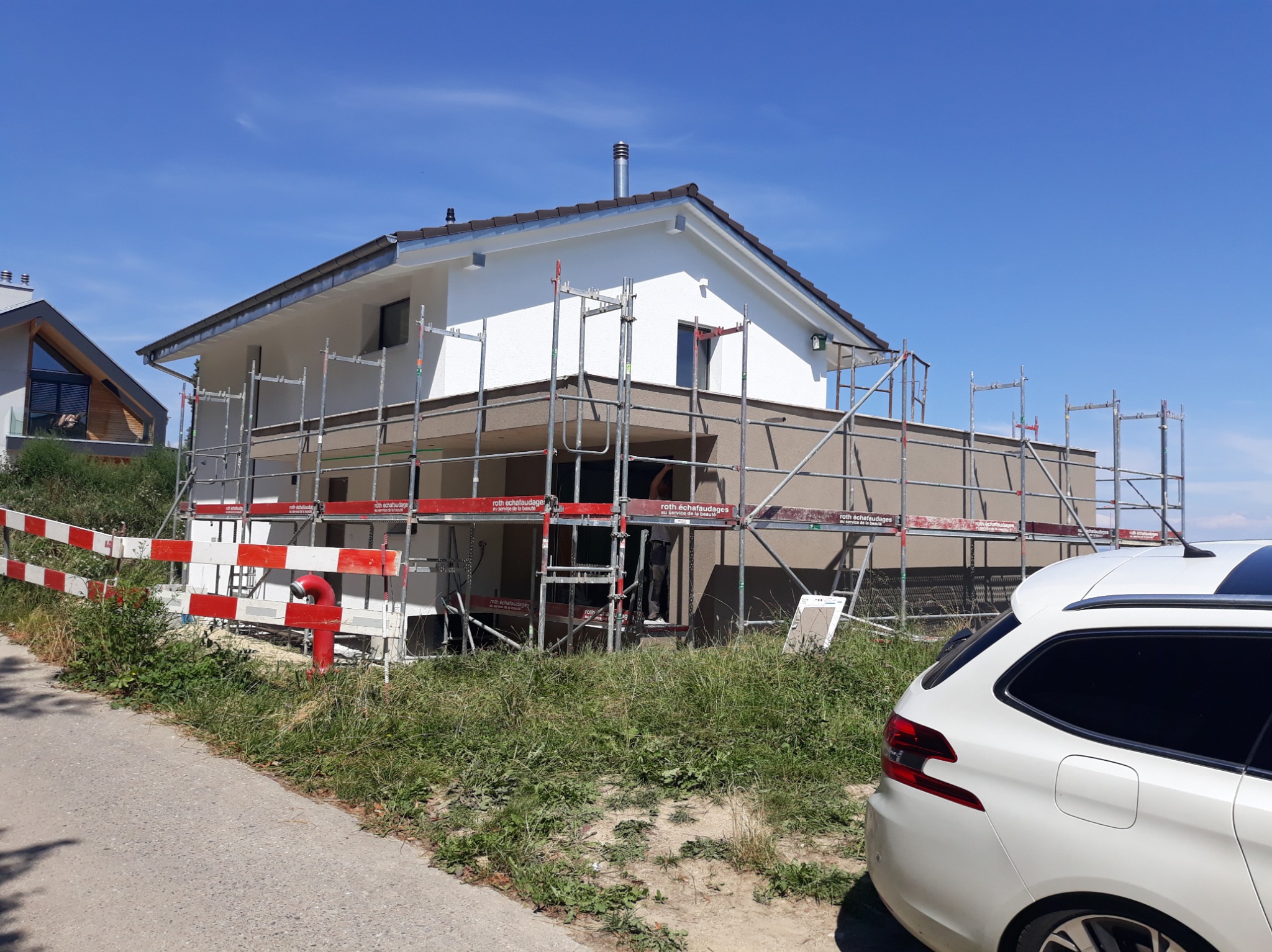 Dom jednorodzinny w trakcie prac elewacyjnych, z widocznymi rusztowaniami i częściowo otynkowaną ścianą. Na pierwszym planie fragment zaparkowanego samochodu.