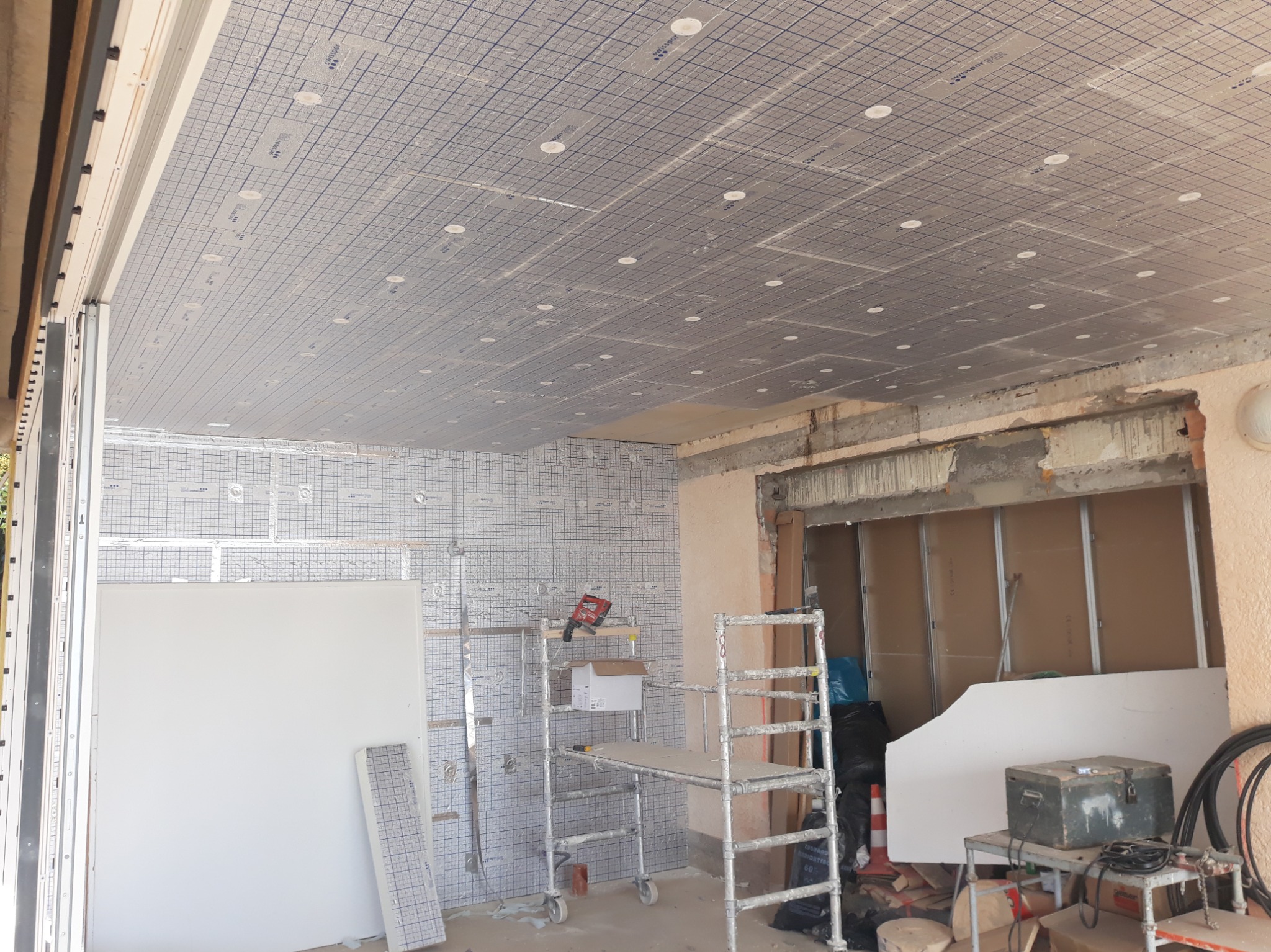 Wnętrze w trakcie prac wykończeniowych: ocieplony sufit z widoczną siatką, rusztowanie, płyty gipsowe. Elementy instalacji widoczne na ścianach.