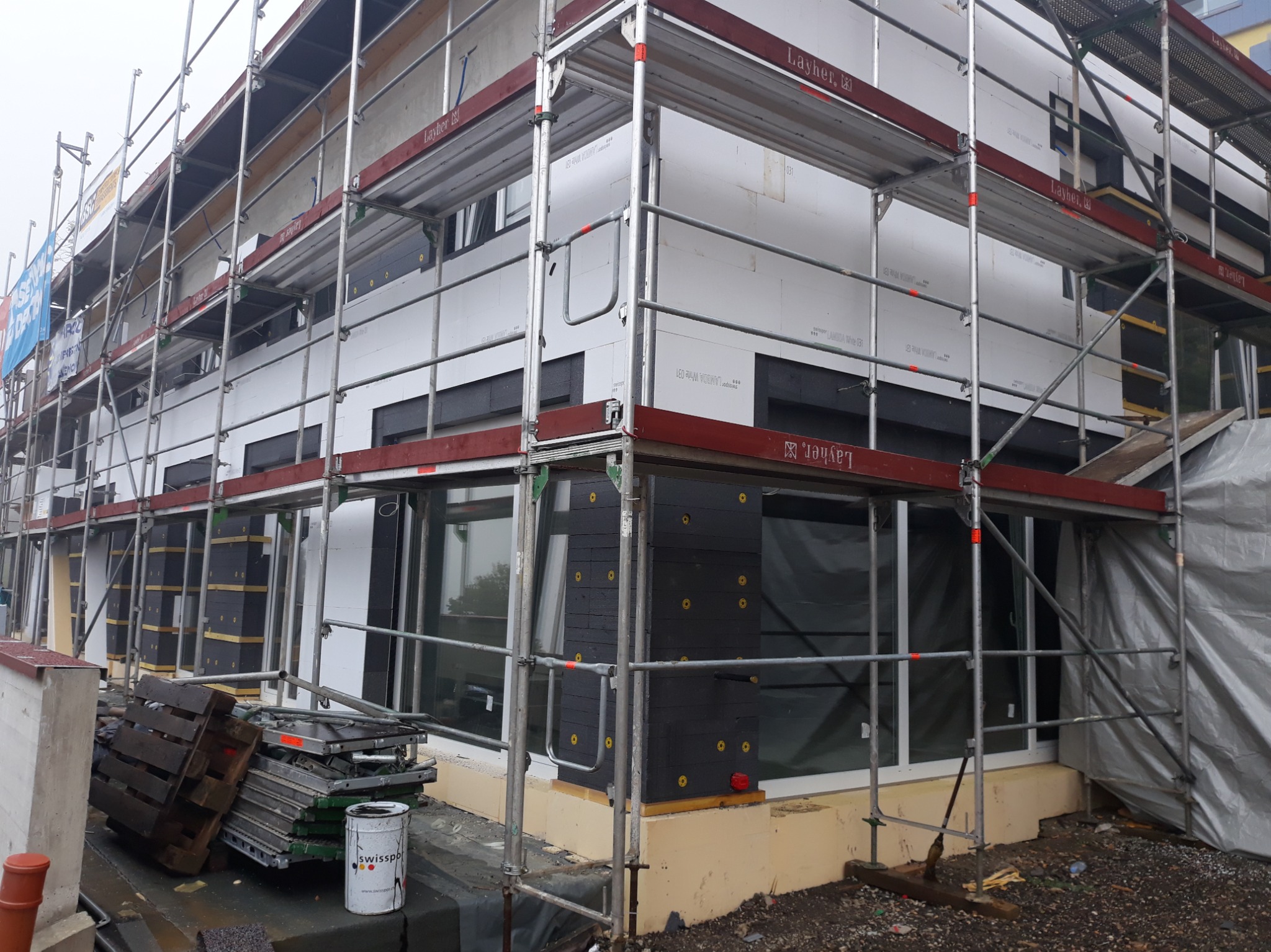 Budynek w trakcie ocieplania, widoczne rusztowania i styropianowe płyty. Część elewacji wykończona ciemnymi panelami.  Na ziemi paleta i puszka farby.