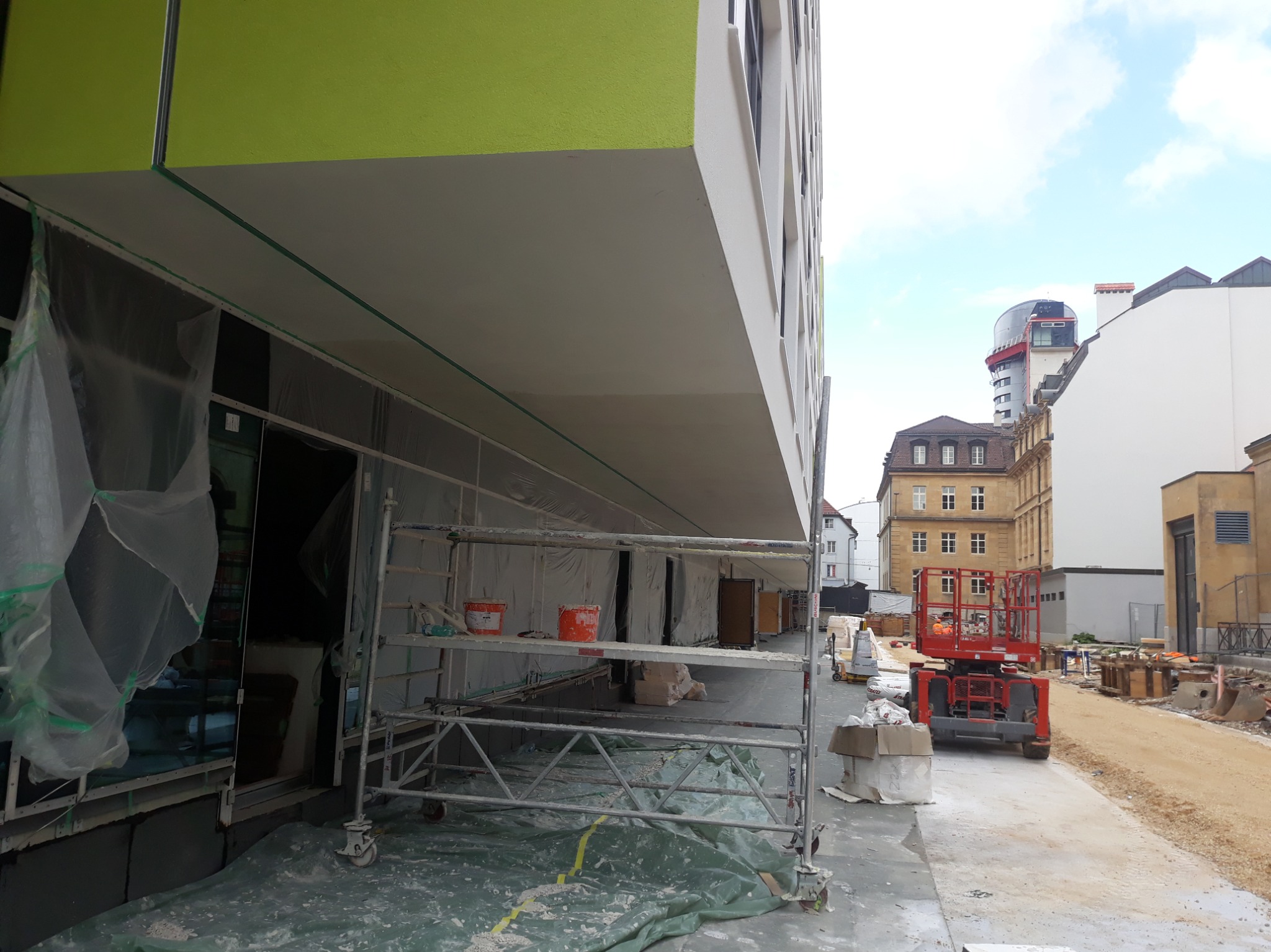 Elewacja budynku w trakcie prac wykończeniowych: rusztowania, zabezpieczenia folią, widoczne puszki z farbą. W tle zabudowa miejska i podnośnik koszowy.