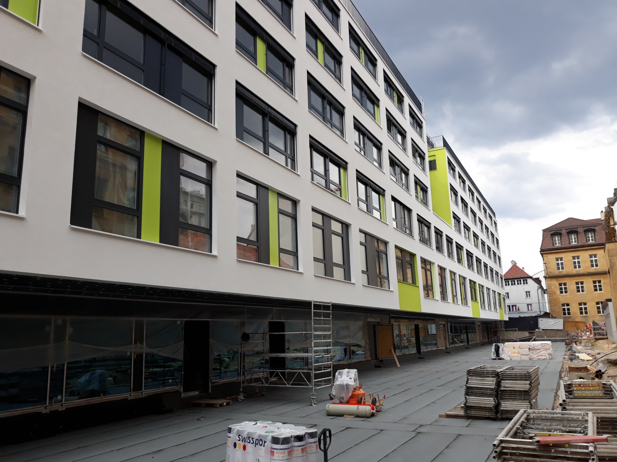 Nowoczesna, biała elewacja budynku z zielonymi akcentami, widoczne rusztowania i materiały budowlane, sugerujące trwające prace wykończeniowe.