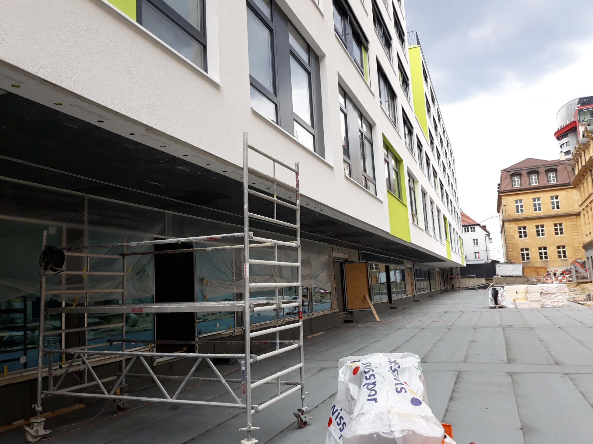 Nowoczesny budynek z białymi ścianami i zielonymi akcentami, widoczny z rusztowaniem i materiałami budowlanymi. Elewacja w trakcie prac wykończeniowych.