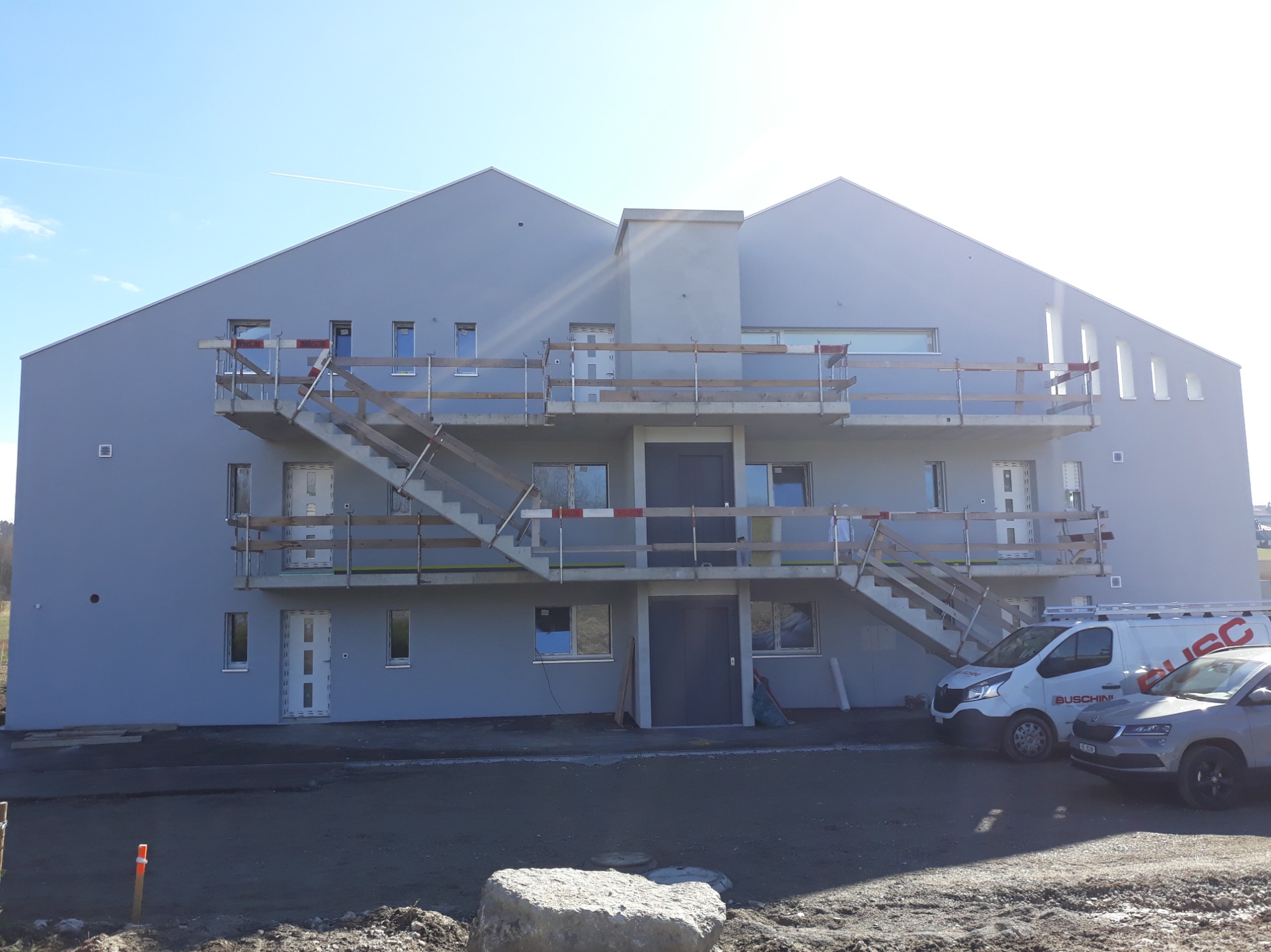 Szara elewacja budynku wielorodzinnego z zewnętrznymi schodami, częściowo osłonięta rusztowaniem, w słoneczny dzień. W tle zaparkowane samochody.
