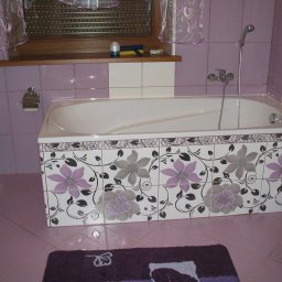 F.H.MIKA MONIKA ZDANCEWICZ - Biała wanna obudowana płytkami z motywem kwiatowym w odcieniach fioletu i szarości, fioletowe płytki ścienne i podłogowe, zasłonka w oknie, widoczna toaleta i dywanik w podobnej kolorystyce.