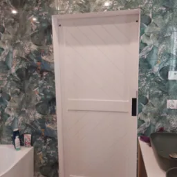 Białe, przesuwne drzwi do łazienki z ukośnymi panelami na tle ściany wyłożonej płytkami z motywem tropikalnych ptaków i roślin, widoczna umywalka i fragment wanny.