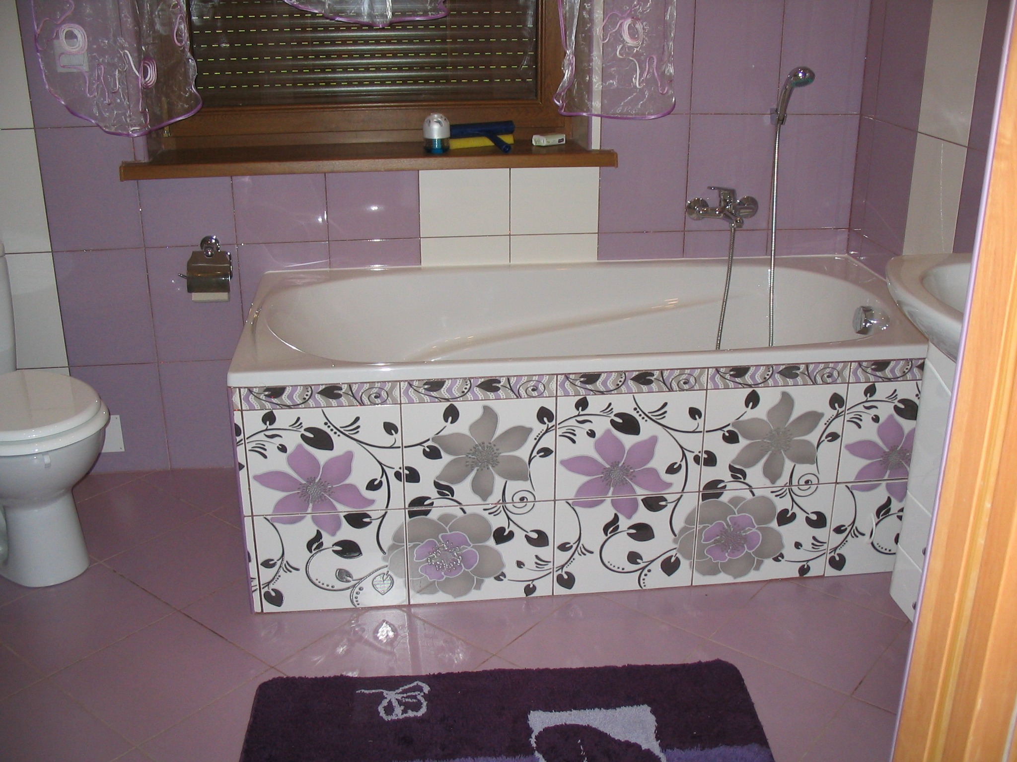 Biała wanna obudowana płytkami z motywem kwiatowym w odcieniach fioletu i szarości, fioletowe płytki ścienne i podłogowe, zasłonka w oknie, widoczna toaleta i dywanik w podobnej kolorystyce.