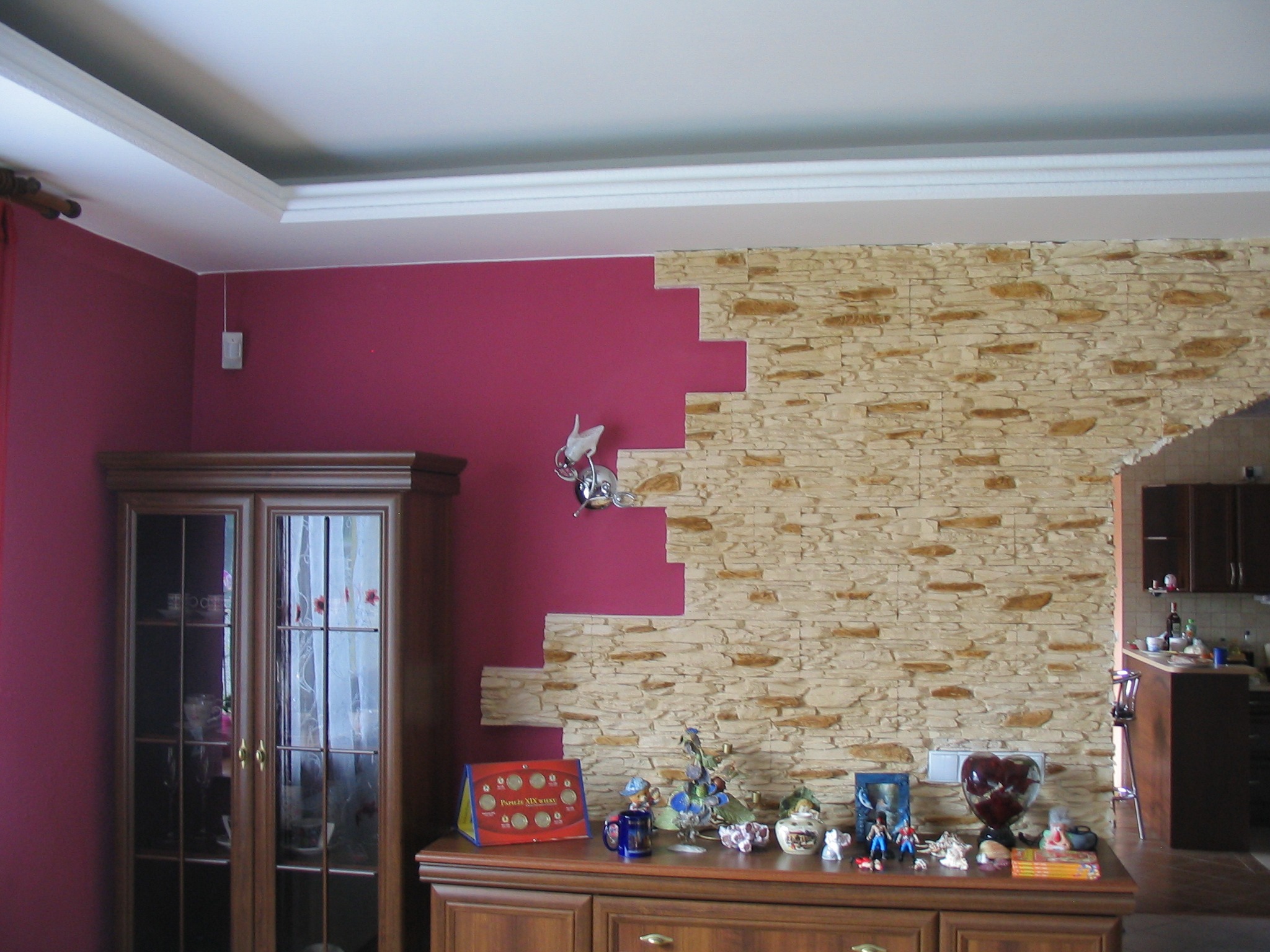 Fragment ściany w salonie z częściowo położoną dekoracyjną okładziną imitującą kamień, przylegającą do ściany pomalowanej na intensywny różowy kolor, z widocznym drewnianym kredensem i dekoracjami.