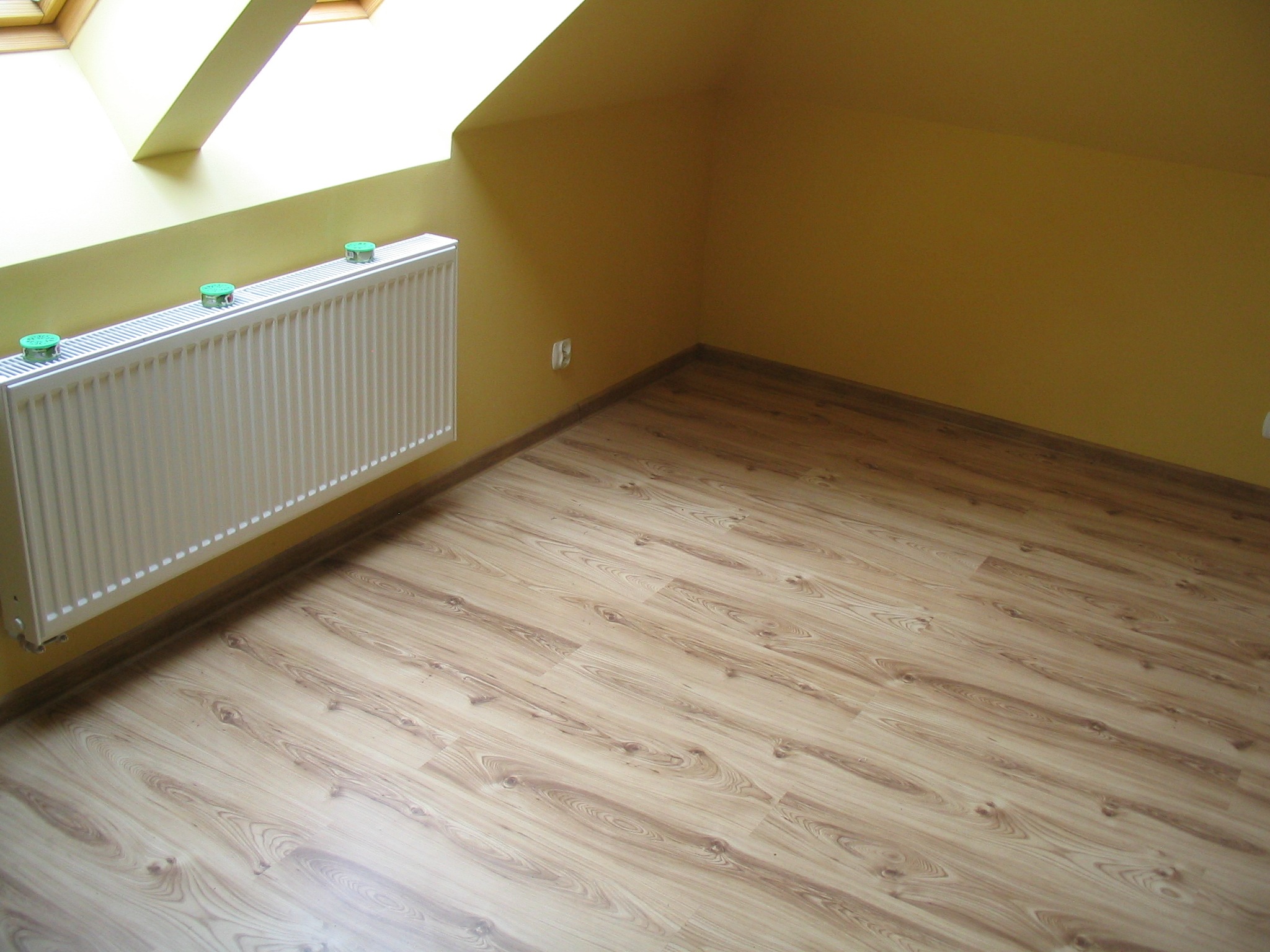 Poddasze po remoncie z panelami podłogowymi imitującymi drewno, żółtymi ścianami i białym grzejnikiem z zielonymi odpowietrznikami, oświetlone światłem wpadającym przez okno dachowe.