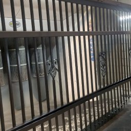 Victoria Lak Pro - Malowanie proszkowe metalowego ogrodzenia z ozdobnymi elementami przez pracownika w kombinezonie ochronnym i masce.