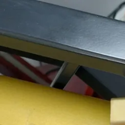 Fragment konstrukcji metalowej: czarna, matowa rura kwadratowa nad złotą, okrągłą rurą, z widocznymi elementami mocującymi i tłem w nieostrym ujęciu.