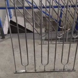 Fragment balustrady ze stali nierdzewnej z pionowymi prętami o zaokrąglonym kształcie u dołu, widoczne otwory montażowe w dolnej listwie, w tle fragment schodów z niebieską balustradą.