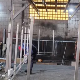 Spawanie konstrukcji ze stali nierdzewnej w warsztacie, widoczne elementy balustrady i spawacz w trakcie pracy, ceglana ściana w tle.