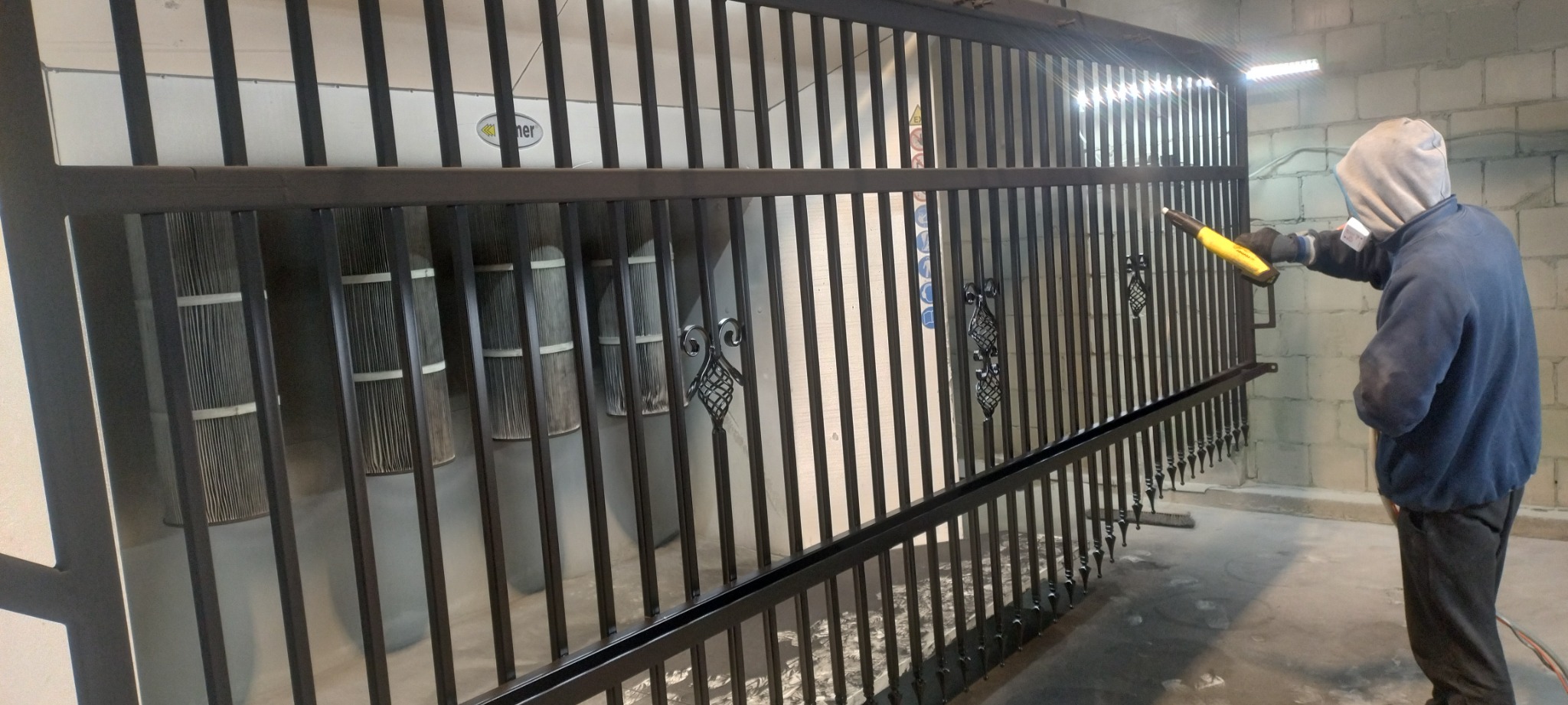 Malowanie proszkowe metalowego ogrodzenia z ozdobnymi elementami przez pracownika w kombinezonie ochronnym i masce.