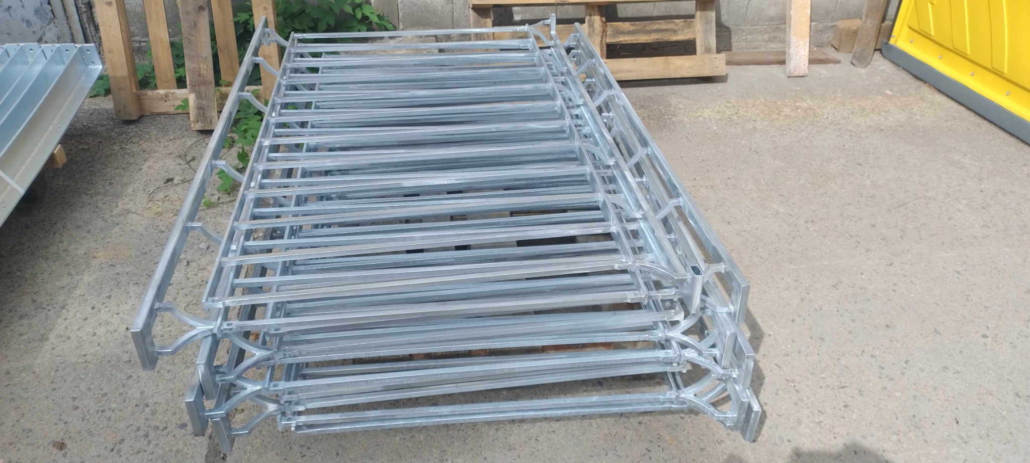 Stos ocynkowanych, metalowych ram z dekoracyjnymi elementami, prawdopodobnie do ogrodzenia, leżący na betonowym podłożu w pobliżu drewnianych palet i żółtej kabiny.