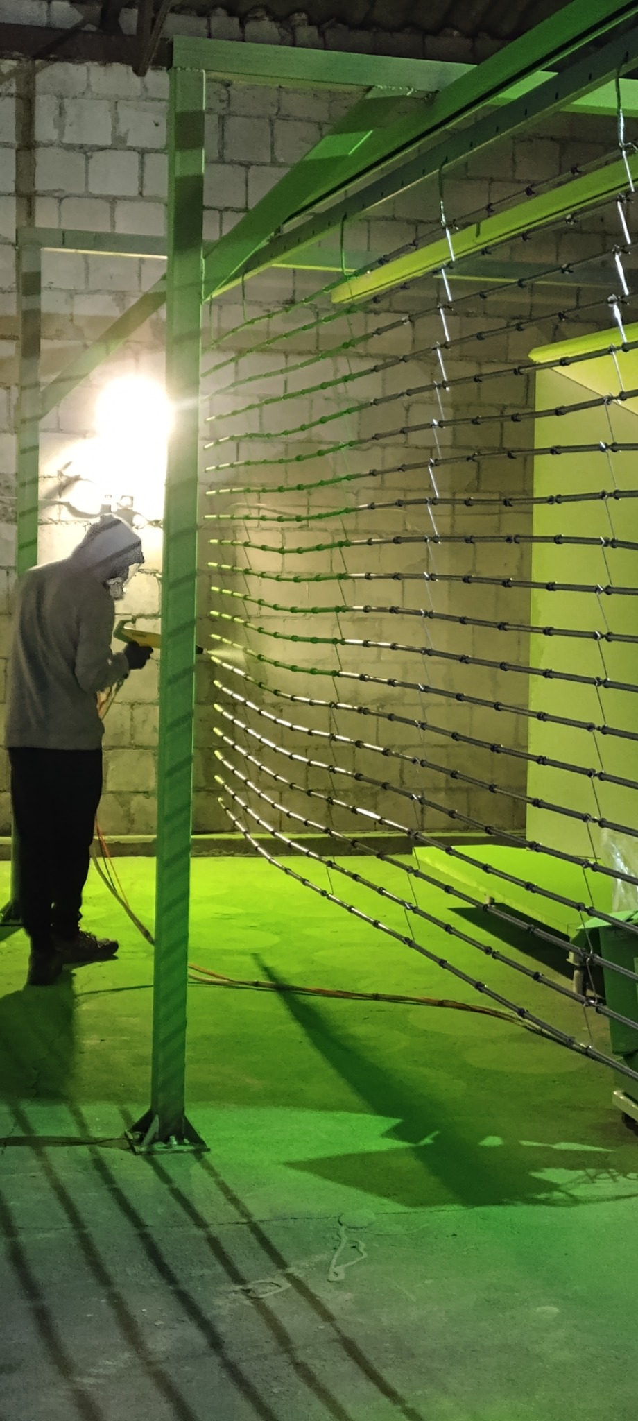 Pracownik w kombinezonie ochronnym maluje proszkowo metalową konstrukcję w hali produkcyjnej, widoczne elementy rusztowania i ceglany mur w tle.