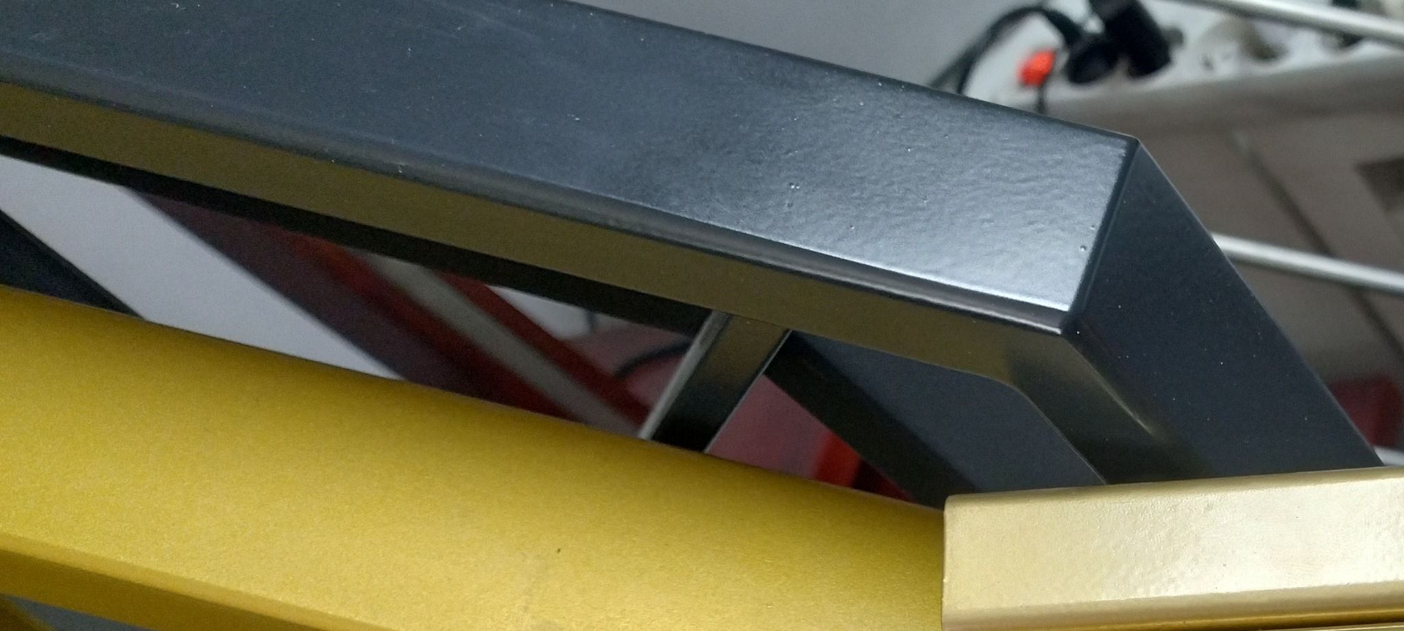 Fragment konstrukcji metalowej: czarna, matowa rura kwadratowa nad złotą, okrągłą rurą, z widocznymi elementami mocującymi i tłem w nieostrym ujęciu.