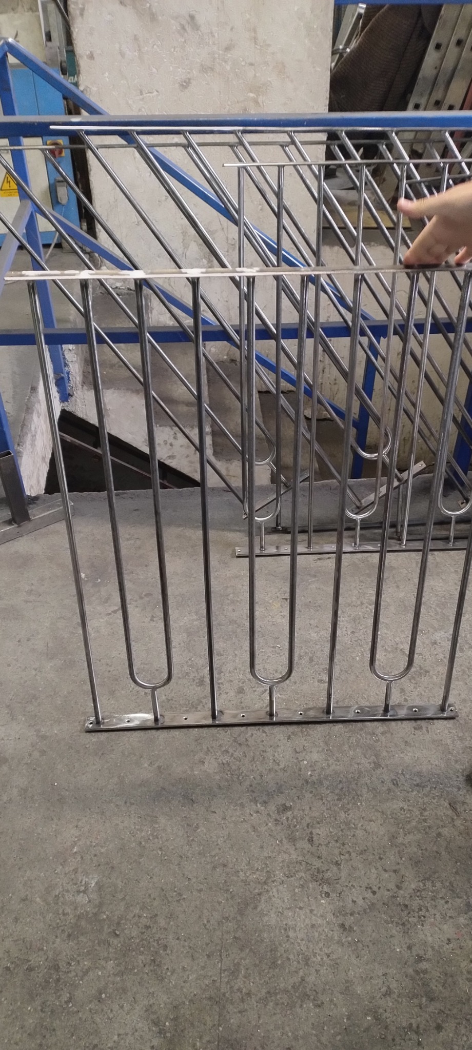 Fragment balustrady ze stali nierdzewnej z pionowymi prętami o zaokrąglonym kształcie u dołu, widoczne otwory montażowe w dolnej listwie, w tle fragment schodów z niebieską balustradą.