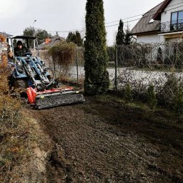MAGPOL Magdalena Polańska - Operator na niebieskim traktorze z glebogryzarką wyrównuje teren pod przyszły trawnik, widoczne przygotowane podłoże i ogrodzenie posesji.