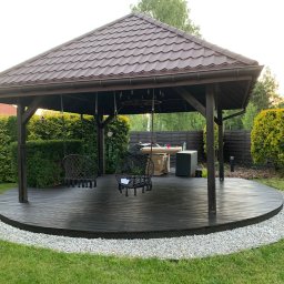 MAGPOL Magdalena Polańska - Drewniana altana z brązowym dachem krytym dachówką, okrągłym tarasem z desek i wiszącymi huśtawkami, otoczona zielonym trawnikiem i krzewami.