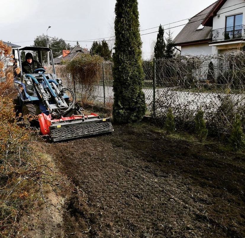 Operator na niebieskim traktorze z glebogryzarką wyrównuje teren pod przyszły trawnik, widoczne przygotowane podłoże i ogrodzenie posesji.