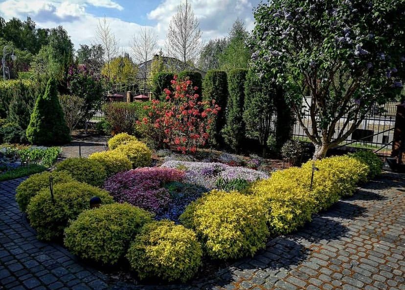 Urokliwy ogród z kwitnącymi rabatami i równo przyciętymi krzewami, otoczony brukowaną ścieżką i wysokimi drzewami, tworzący harmonijną kompozycję.