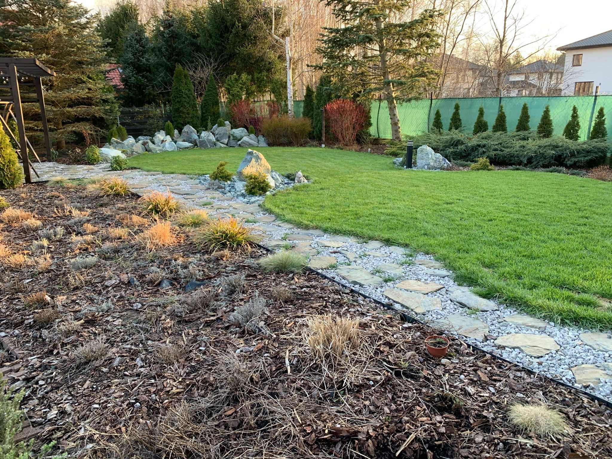 Ułożony kamienny chodnik wijący się przez ogród z równo przyciętym trawnikiem i rabatami z korą, ozdobiony kamieniami i niskimi krzewami. W tle zielona siatka ogrodzeniowa i drzewa.