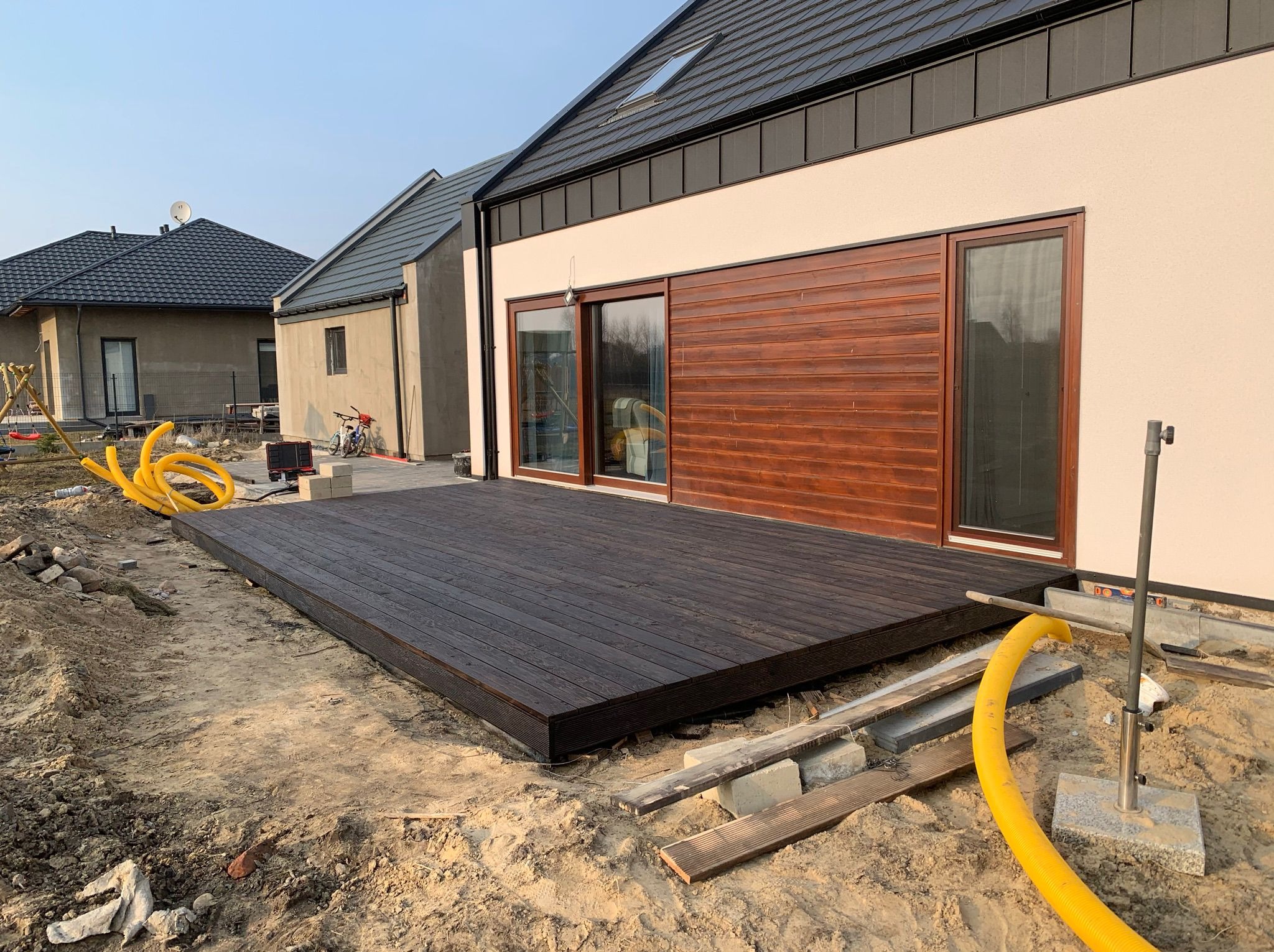 Nowo wybudowany, ciemny taras drewniany przylegający do domu z brązowymi panelami, widoczne żółte rury i elementy konstrukcyjne w surowym otoczeniu.