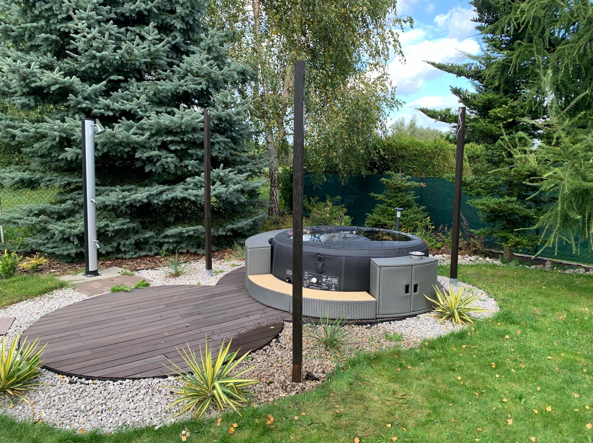 Okrągła, zewnętrzna wanna z hydromasażem na drewnianym podeście, otoczona ozdobnym żwirem i roślinami, z widocznymi elementami instalacji prysznicowej i wysokimi drzewami iglastymi w tle.