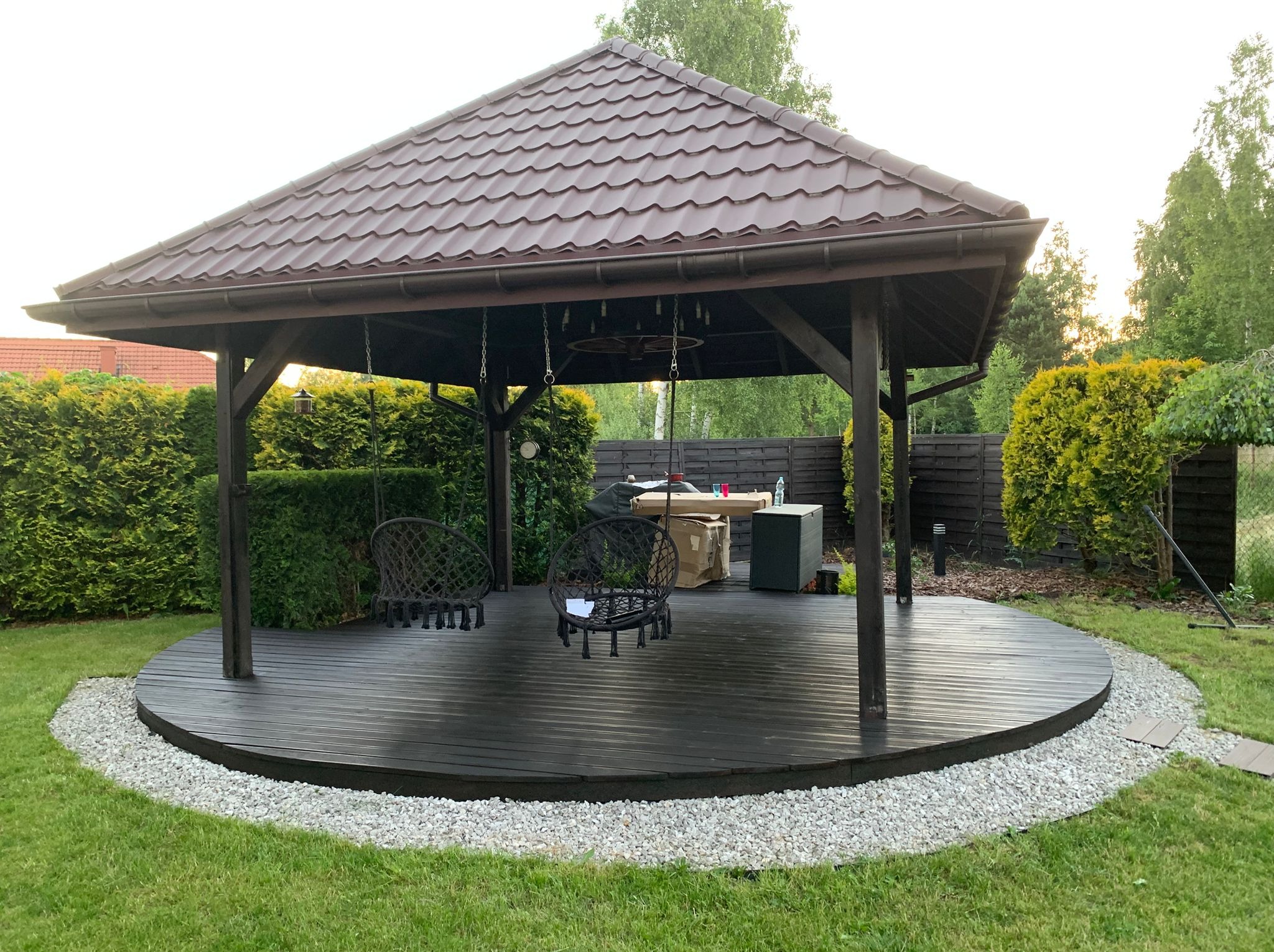 Drewniana altana z brązowym dachem krytym dachówką, okrągłym tarasem z desek i wiszącymi huśtawkami, otoczona zielonym trawnikiem i krzewami.