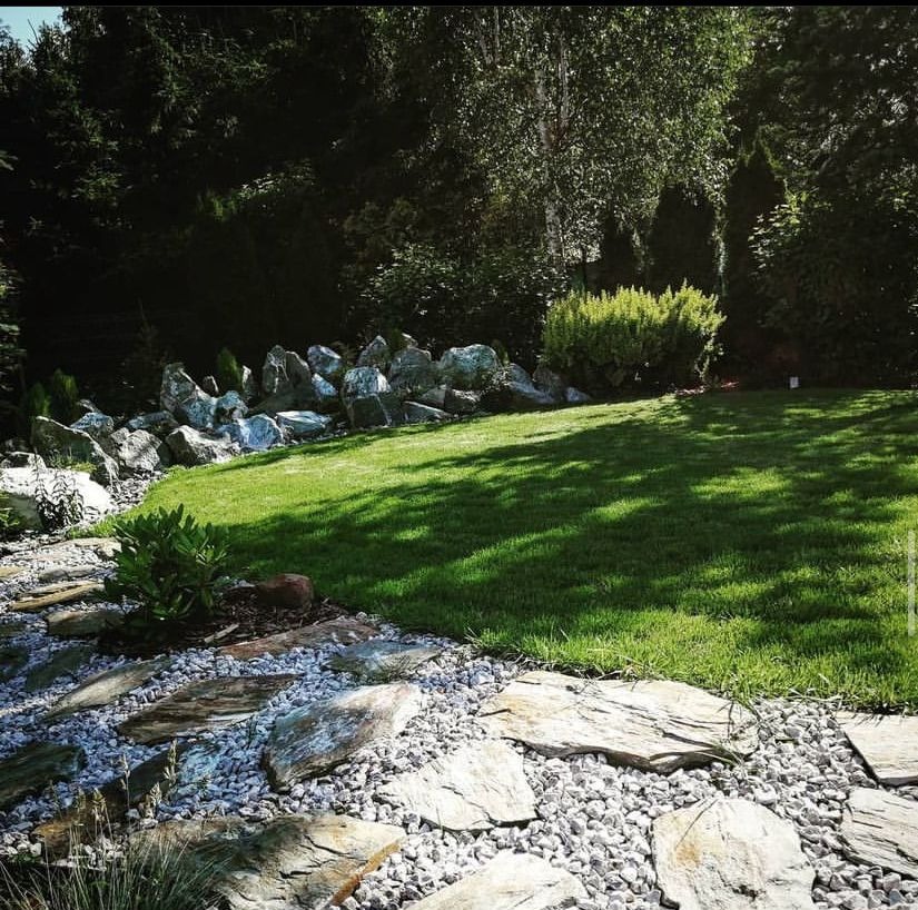 Ujęcie ogrodu z równo przystrzyżonym trawnikiem, ścieżką z nieregularnych kamieni i otaczającymi roślinami oraz dekoracyjnym skalniakiem z dużych kamieni w tle.