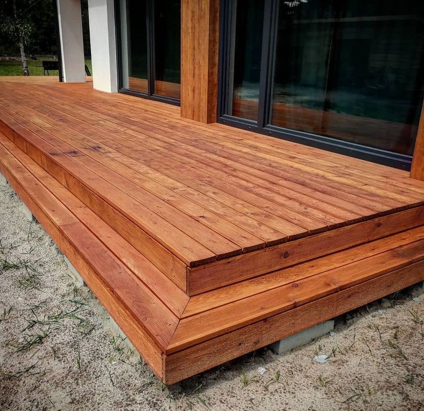 Drewniany taras z schodami wykonany z desek w odcieniach brązu, przylegający do budynku z dużymi oknami.