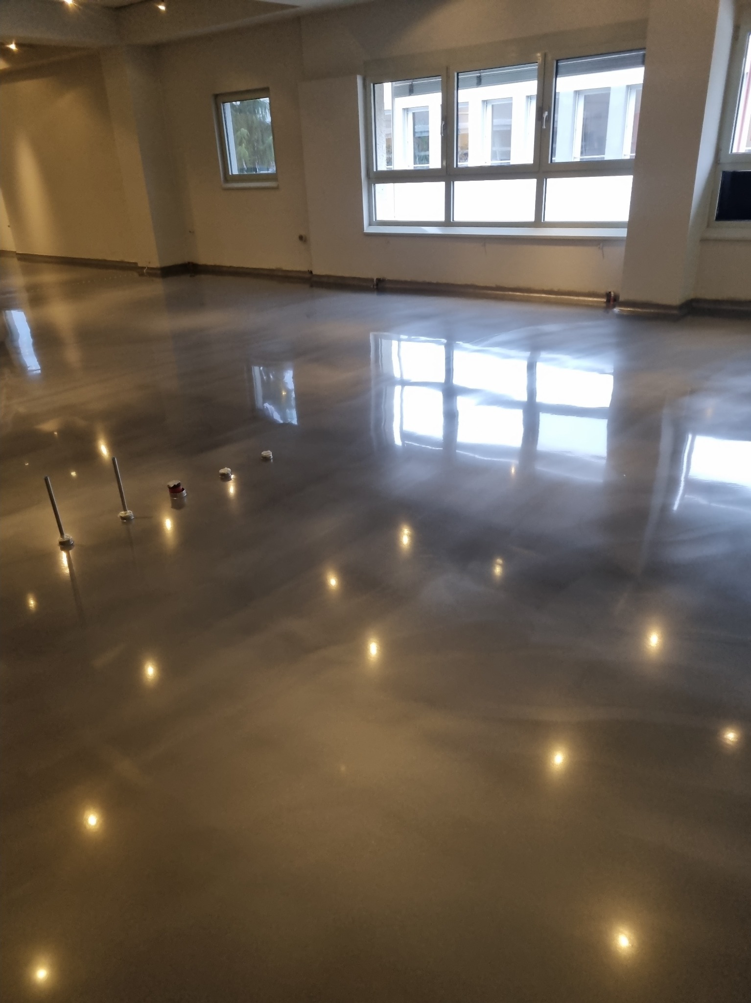 Błyszcząca, szara posadzka w przestronnym, jasnym pomieszczeniu z dużymi oknami, widoczne narzędzia i akcesoria do montażu w trakcie prac.