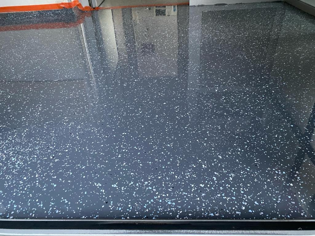 Błyszcząca, ciemnoszara posadzka z drobnymi, srebrnymi wtrąceniami, świeżo wylana, z zabezpieczeniem przy ścianie.