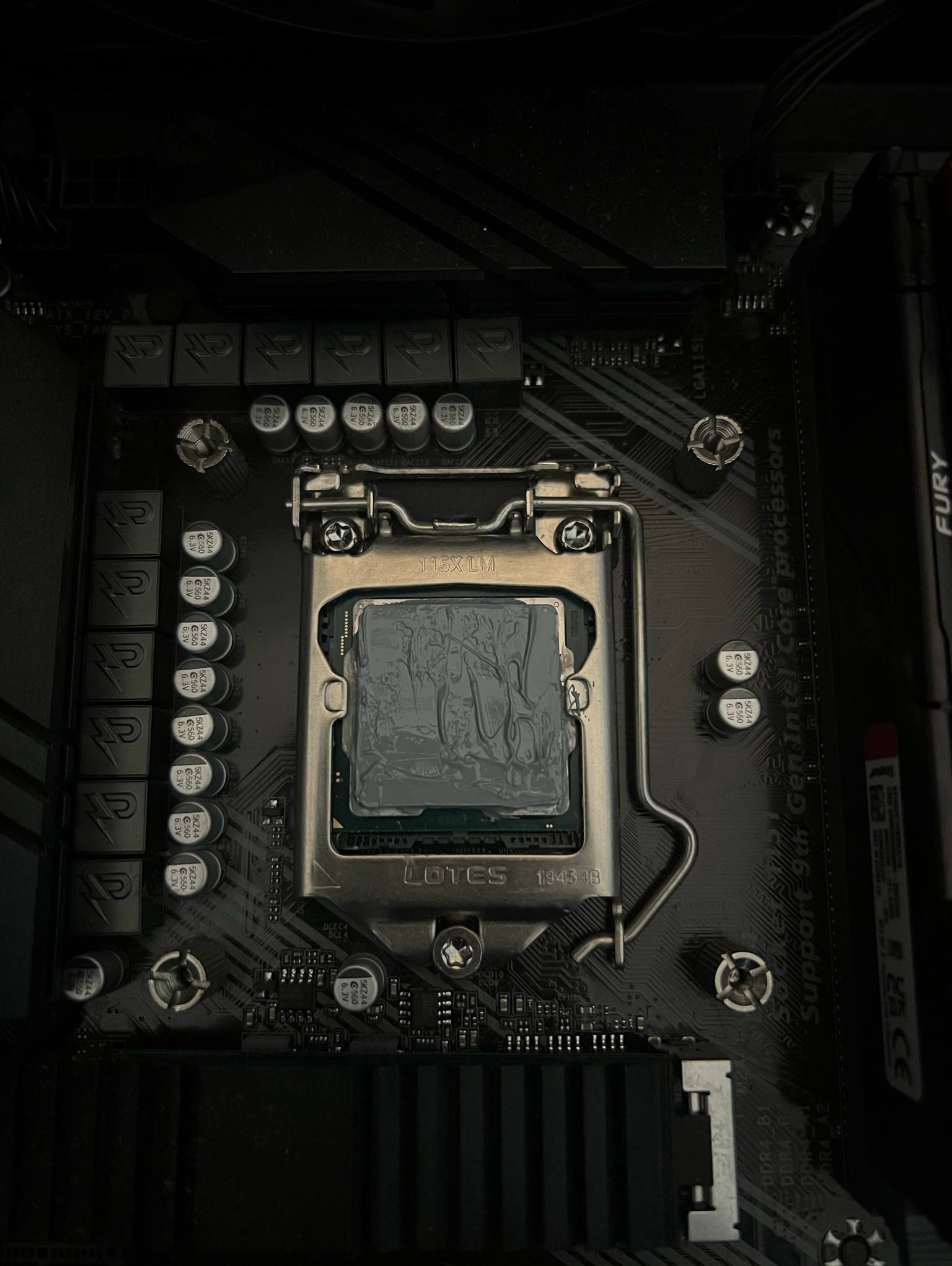 Płyta główna z zamontowanym procesorem Intel, pokrytym pastą termoprzewodzącą, gotowa do montażu chłodzenia. Widoczne elementy płyty głównej i gniazdo procesora.