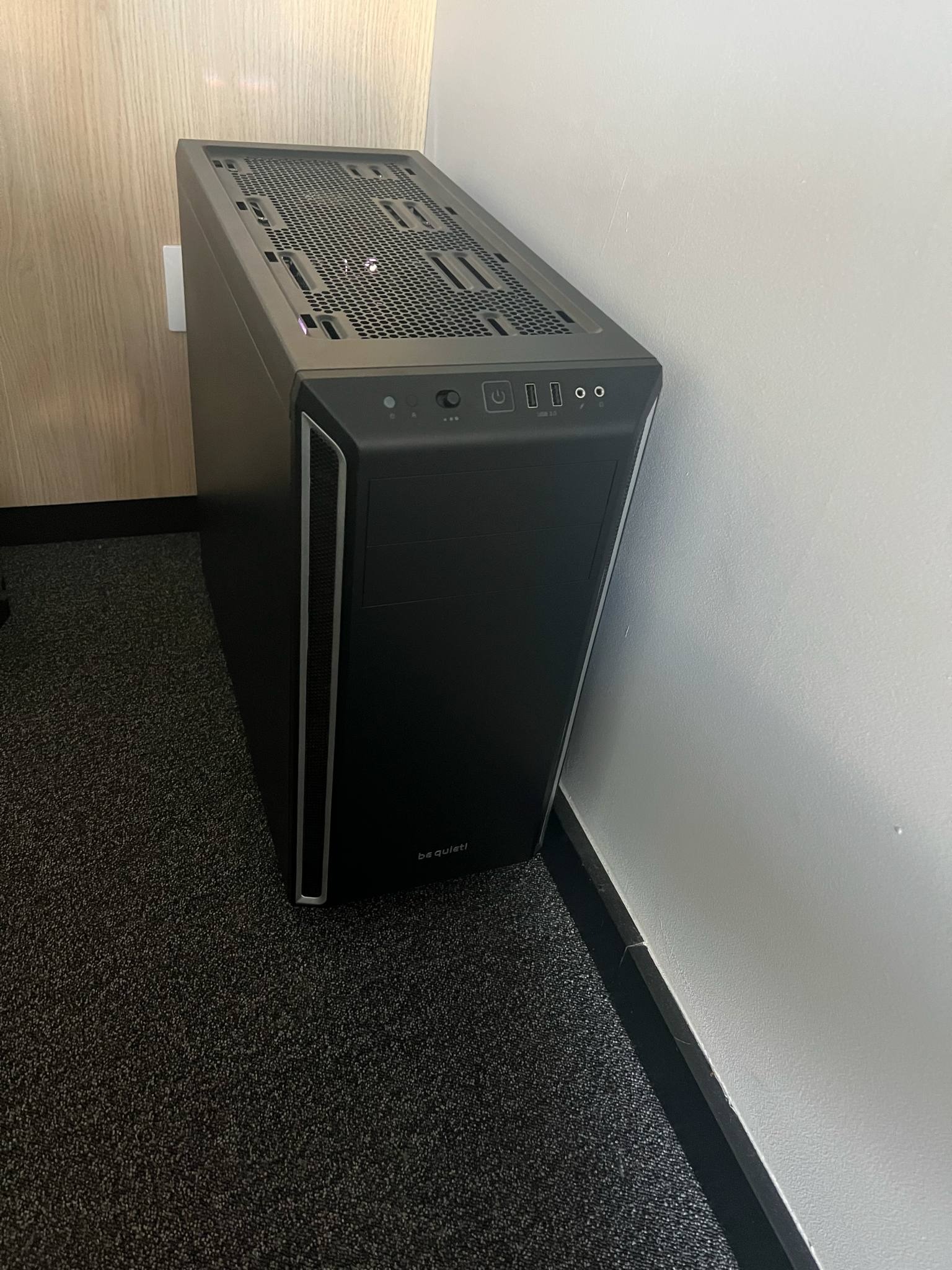 Czarna obudowa komputera stacjonarnego marki be quiet! ustawiona pionowo na ciemnej wykładzinie, widoczne porty USB i przycisk zasilania na górnym panelu.
