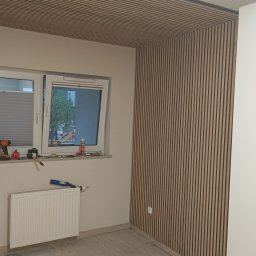 Kam.gru.bud. - Montaż paneli ściennych i sufitowych z lameli w pokoju z oknem, widoczne narzędzia pracy i grzejnik.