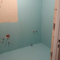 Kam.gru.bud. - Świeżo pomalowana na turkusowo łazienka w trakcie prac wykończeniowych, widoczne wystające rury i kable, fragment ściany w kolorze beżowym z niedomalowanym obszarem, surowe obramowanie drzwi.