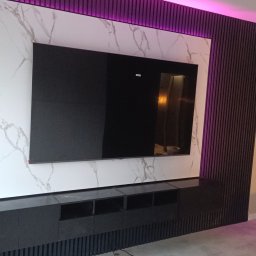 Kam.gru.bud. - Nowoczesna ściana telewizyjna z podświetleniem LED, wykończona panelami z ciemnego drewna i marmurową płytą, z czarną szafką RTV na dole.