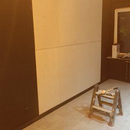 Kam.gru.bud. - Ściana w trakcie remontu: fragment pomalowany na ciemny brąz, reszta w jasnych płytach, drabina z narzędziami malarskimi w pomieszczeniu z szarą podłogą imitującą beton.